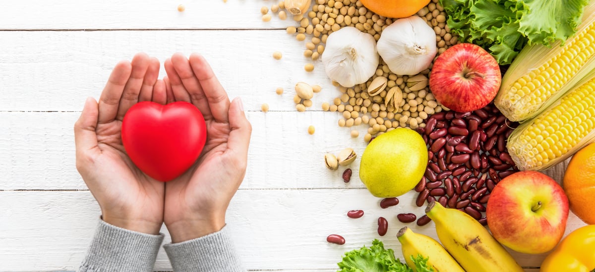 White Hands Holding a Plastic Heart Beside a Pile of Healthy Food